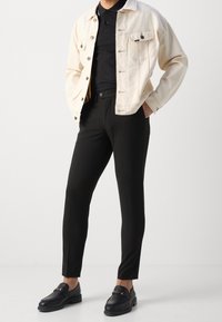 Licht crème denim jacket over een zwart polo, gecombineerd met op maat gemaakte zwarte broek en zwarte loafers, met een decoratieve gouden accent.