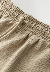 Pantalon en tissu beige texturé avec une taille élastique et un cordon de serrage noué à l'avant.