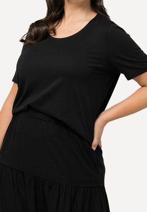 Femme portant un haut noir ample à manches courtes et une jupe, une main reposant sur sa hanche sur un fond uni.