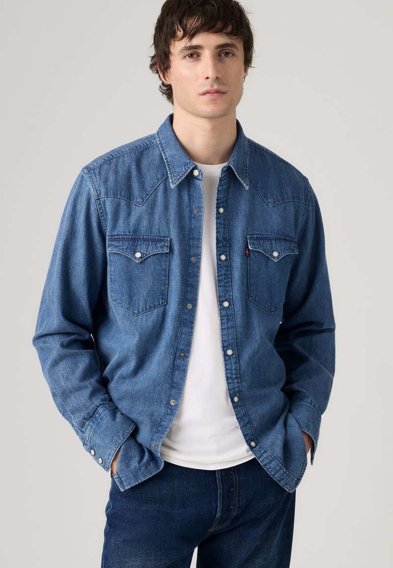Jeune homme portant une chemise en denim bleu sur un t-shirt blanc, les mains dans les poches, sur fond uni.