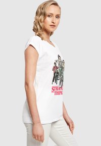 Valge puuvillane T-särk, ümmarguse kaelusega, lühikeste varrukatega, eespool erksate värvidega "Stranger Things" graafilise kujutisega.