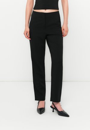 Donna che indossa pantaloni dritti a vita alta neri, top corto nero e décolleté a punta nere su uno sfondo bianco.