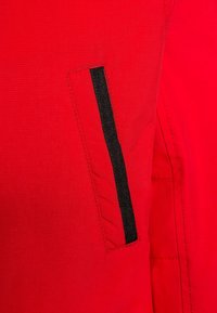 Detalje af en rød stofjakke med en lomme med en sort lodret stribe akcent, glat tekstur og ren syning.