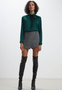 Blusa verde in satin con colletto annodato, abbinata a una gonna avvolgente in tessuto a fantasia cavalli bianchi e neri e stivali in pelle neri alti fino alla coscia.