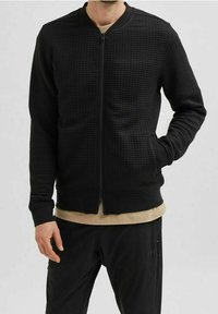 Černá bomber bunda s textured látkou, zapínáním na zip, ribovanými manžetami a lemem; v kombinaci s béžovým tričkem a černými kalhotami.