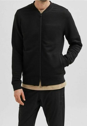 Giacca bomber nera in tessuto testurizzato, con chiusura a zip, polsini e orlo a costine; abbinata a una maglietta beige e pantaloni neri.