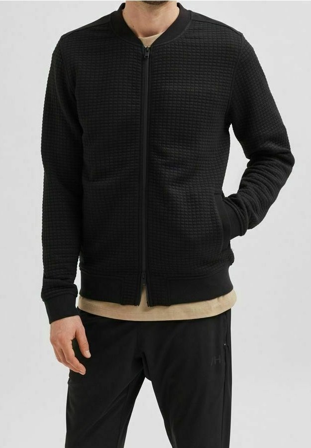 Černá bomber bunda s textured látkou, zapínáním na zip, ribovanými manžetami a lemem; v kombinaci s béžovým tričkem a černými kalhotami.