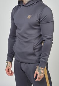 Felpa grigia scura realizzata in un materiale liscio e elastico. Presenta una tasca frontale, un logo dorato e pantaloni abbinati con una striscia dorata laterale.