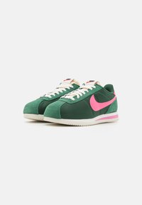 Zelené semišové a textilní tenisky s růžovým logo Nike, smetanovými tkaničkami a bílou gumovou podrážkou, s zaoblenou špičkou a nízkým profilem.