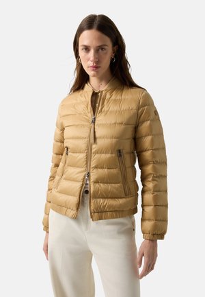 Kobieta nosząca beżową pikowaną kurtkę zapinaną na zamek z kieszeniami na zamek, w połączeniu z jasnobeżowymi spodniami z wysokim stanem, stojąca na jednolitym tle.