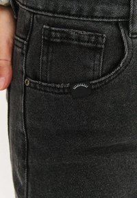 Čierne rifle z denimu s predným vreckom, na ktorom je malá čierna etiketa so svetlým grafickým znázornením zakrivených mihalníc. Hladká, textúrovaná látka.