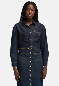 Chaqueta de mezclilla con mangas largas y un bolsillo frontal, combinada con una falda de mezclilla oscura que presenta botones plateados y dos bolsillos frontales.