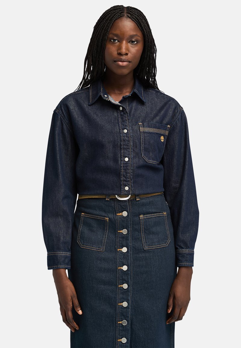 Chaqueta de mezclilla con mangas largas y un bolsillo frontal, combinada con una falda de mezclilla oscura que presenta botones plateados y dos bolsillos frontales.