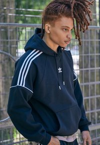 Felpa blu navy con strisce bianche sulle maniche, cappuccio regolabile con lacci, tasca frontale e piccolo logo Adidas al centro del petto.