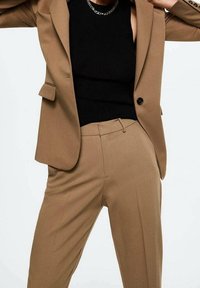 Personne portant un costume couleur camel avec un blazer à un bouton sur un haut sans manches noir et un collier en chaîne argenté.