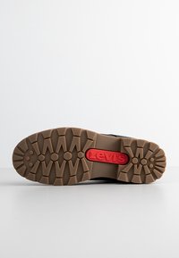 Semelle de chaussure en caoutchouc marron avec des rainures profondes, présentant un logo Levi's rouge au centre, affichée sur un fond blanc uni.