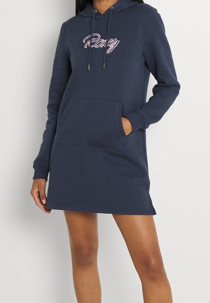 Vestido sudadera azul marino de tejido suave con bolsillo frontal. Presenta el logo en rosa "Roxy" y capucha ajustable con cordón. Los puños y el dobladillo son de canalé.