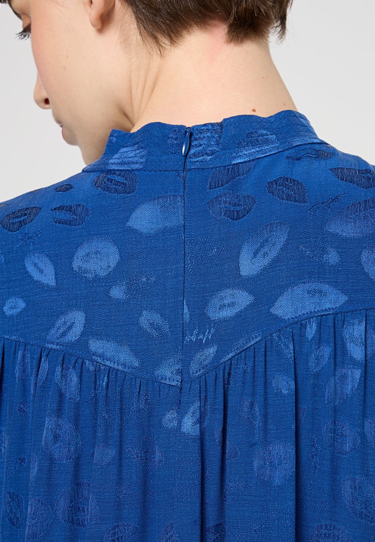 Blaue Bluse aus strukturiertem Stoff mit hohem Kragen, Reißverschluss an der Rückseite und einem abstrakten Lippenmuster. Falten-Details hinten.