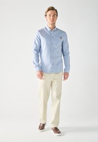 Light blue button-up shirt with a chest pocket, brown buttons, and a leather logo accent, paired with beige trousers and brown shoes.