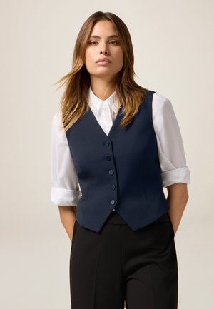 Marineblauer Weste mit fünf Knöpfen, taillierte Passform, V-Ausschnitt, kombiniert mit einem weißen Hemd mit Kragen, präsentiert an einem Modell. Schwarze Hosen runden den Look ab.