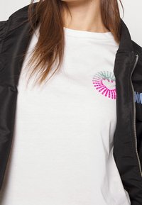Bílý t-shirt s barevným kruhovým potiskem s designem srdce, převalený pod černou bomber bundou. Hladká textura látky.