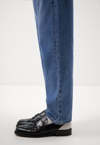 Jambes portant un jean bleu en denim et des mocassins en cuir noir avec des chaussettes de couleur claire sur un fond blanc uni.