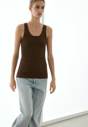 Jeune femme marchant à l'intérieur portant un haut sans manches côtelé marron et un jean bleu clair, avec une expression faciale neutre.