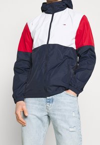 Färgsorterad vindjacka med navyblå kropp, vita och röda ärmar, dragkedja fram och elastiska cuffs. Har en huva och sidfickor.