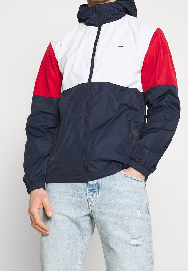 Färgsorterad vindjacka med navyblå kropp, vita och röda ärmar, dragkedja fram och elastiska cuffs. Har en huva och sidfickor.