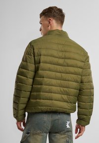 Grön pufferjacka med ribbat mönster och horisontella sömmar. Den har en ståkrage och är kombinerad med blå denimbyxor.