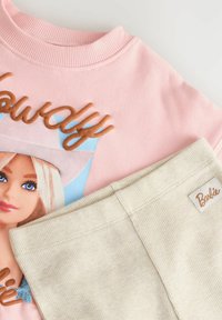 Sweat-shirt rose avec une illustration de Barbie et un pantalon beige texturé avec une petite étiquette Barbie cousue à la taille.