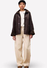 Personne portant une veste en shearling marron foncé, un pantalon large couleur crème, un haut blanc et des chaussures à imprimé léopard, se tenant devant un fond uni.