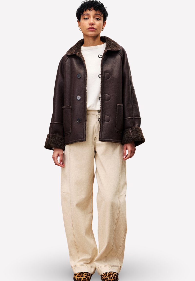 Personne portant une veste en shearling marron foncé, un pantalon large couleur crème, un haut blanc et des chaussures à imprimé léopard, se tenant devant un fond uni.