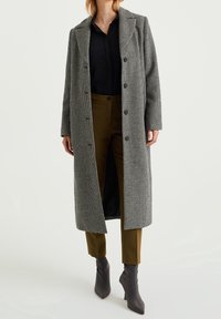 Cappotto lungo con motivo pied de poule in bianco e nero, caratterizzato da un colletto a risvolto, chiusura con bottoni e tasche frontali, abbinato a pantaloni oliva e stivali grigi.