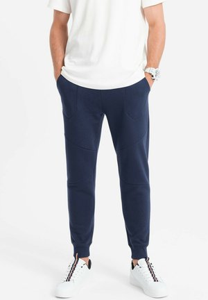 Navy jogger pants made of textured fabric, featuring two front pockets, tapered ankles, and reinforced seams, paired with white sneakers.