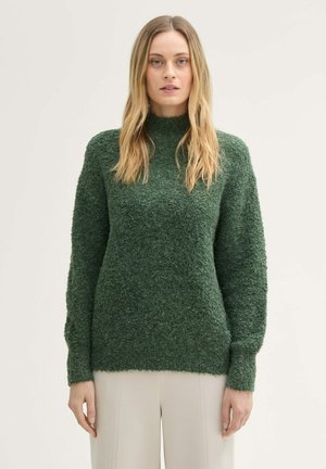 Grüner, strukturierter Strickpullover mit hohem Kragen, langen Ärmeln und gerippten Manschetten, kombiniert mit hellen Hosen.
