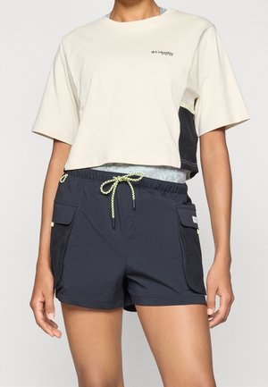 Osoba ubrana w beżowy krótki t-shirt Columbia i czarne spodenki z żółtym sznurkiem i bocznymi kieszeniami na jednolitym tle.
