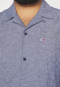 Camicia a maniche corte con colletto in lino blu chiaro, dal tessuto morbido. Presenta una tasca sul petto con un piccolo logo e bottoni scuri.