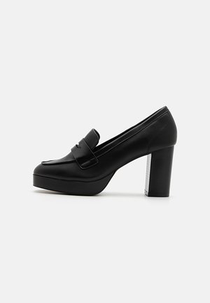 Black leather high-heeled platform loafers with a rounded toe and a decorative strap. The heel features a textured silver accent.