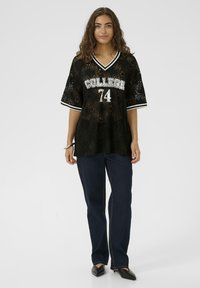 Črna čipkasta majica z grafiko "COLLEGE 74", kratkimi rokavi in V-izrezom; v kombinaciji s temnimi kavbojkami in črnimi čevlji s poudarjenim prednjim delom.