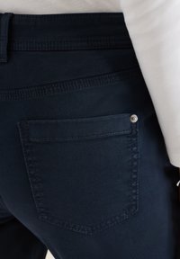 Gros plan sur un pantalon ajusté bleu marine foncé avec une poche arrière visible et des coutures apparentes, porté avec un haut blanc.