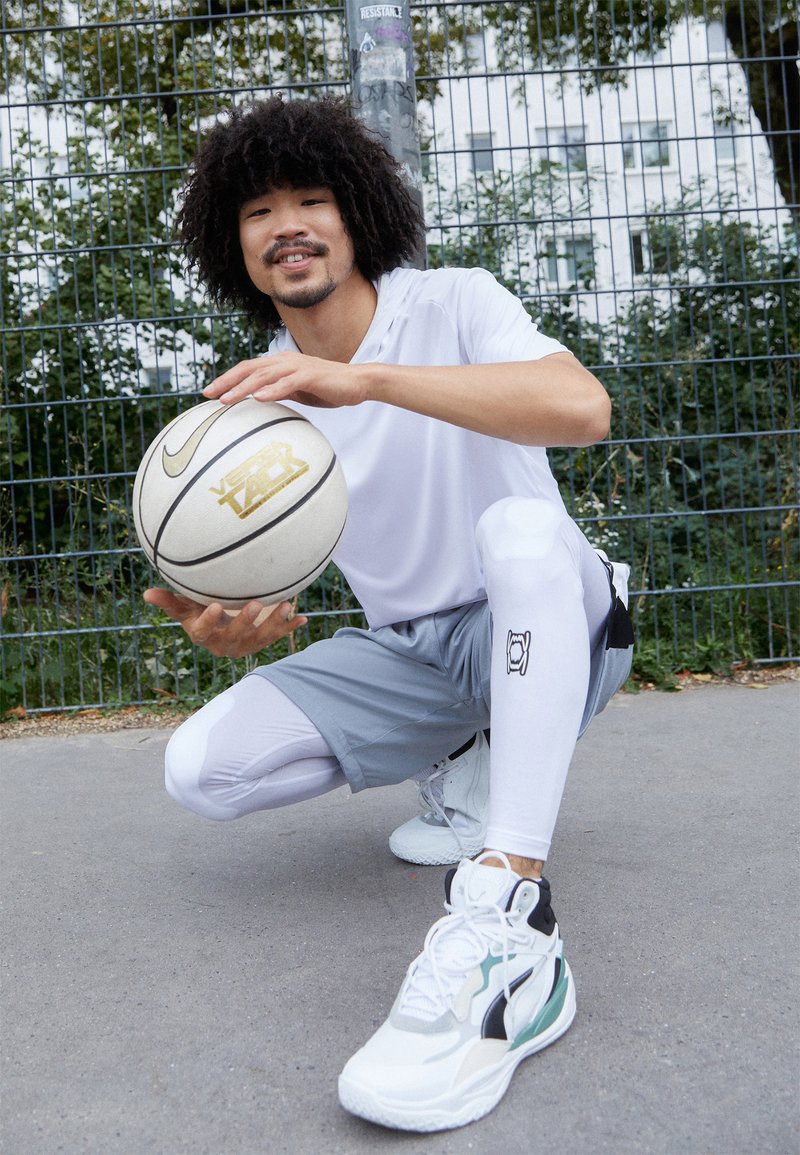 Palla da basket bianca Nike con accenti dorati tenuta da una persona in camicia bianca, pantaloni grigi e scarpe da basket con un design multicolore.