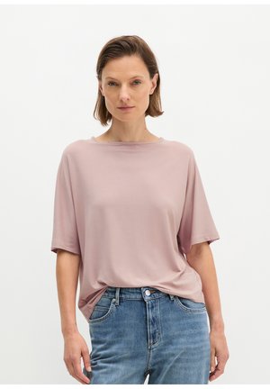 Frau mit kurzen braunen Haaren trägt ein lockeres, hell-mauvefarbenes T-Shirt und blaue Jeans, steht vor einem schlichten weißen Hintergrund.