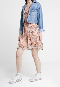 Denimjacke in Hellblau mit hochgekrempelten Ärmeln über einem blumigen Blush-Kleid mit orangefarbenen und grauen Akzenten, kombiniert mit weißen Sneakern.