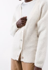 Gestrickte cremefarbene Strickjacke mit einem gerippten Muster, ausgestattet mit einem V-Ausschnitt und sechs schimmernden Knöpfen entlang der Vorderseite. Weicher, warmer Stoff.