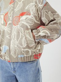 Pletená bomber bunda v béžové barvě s květinovým vzorem v červené, modré a bílé. Má elastické manžety a pas, boční kapsy.