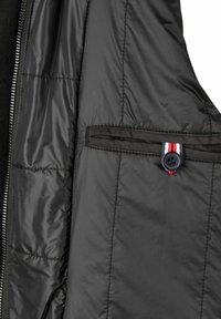 Melnā pūķu jaka ar quilted tekstūru iekšpusē, kas aprīkota ar atloku, kuru droši tur zobens ar sarkanu, baltu un zilu svītru akcentu.