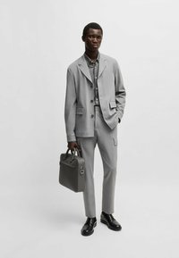 Abito grigio con giacca a un petto, pantaloni abbinati e una valigetta grigia. Tessuto liscio, vestibilità tagliata su misura, design minimalista e scarpe scure.