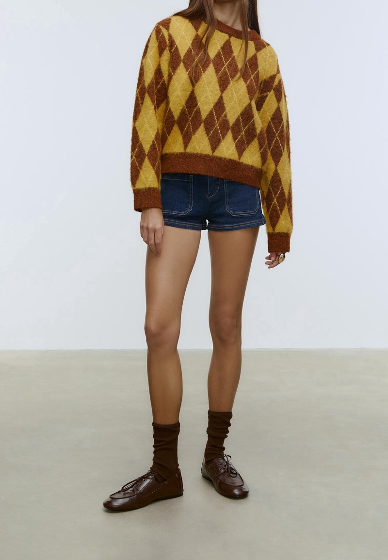 Femme portant un pull jaune et marron à motif losanges, un short en jean bleu foncé, des chaussettes marron et des chaussures en cuir marron à lacets, debout sur un sol gris.