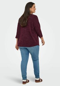 Maglione bordeaux, con texture, a tre quarti indossato sopra un paio di jeans azzurri. L'outfit include scarpe marroni e bianche.